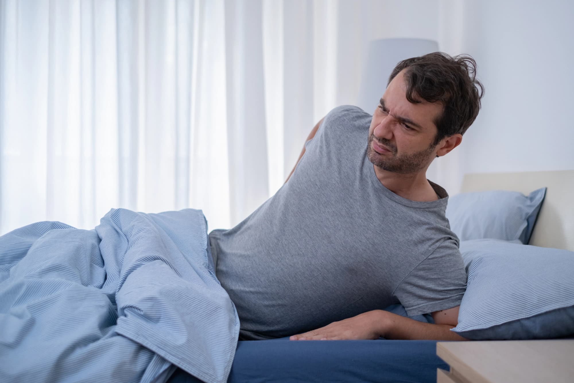 Man in gray shirt lying uncomfortably in bed.