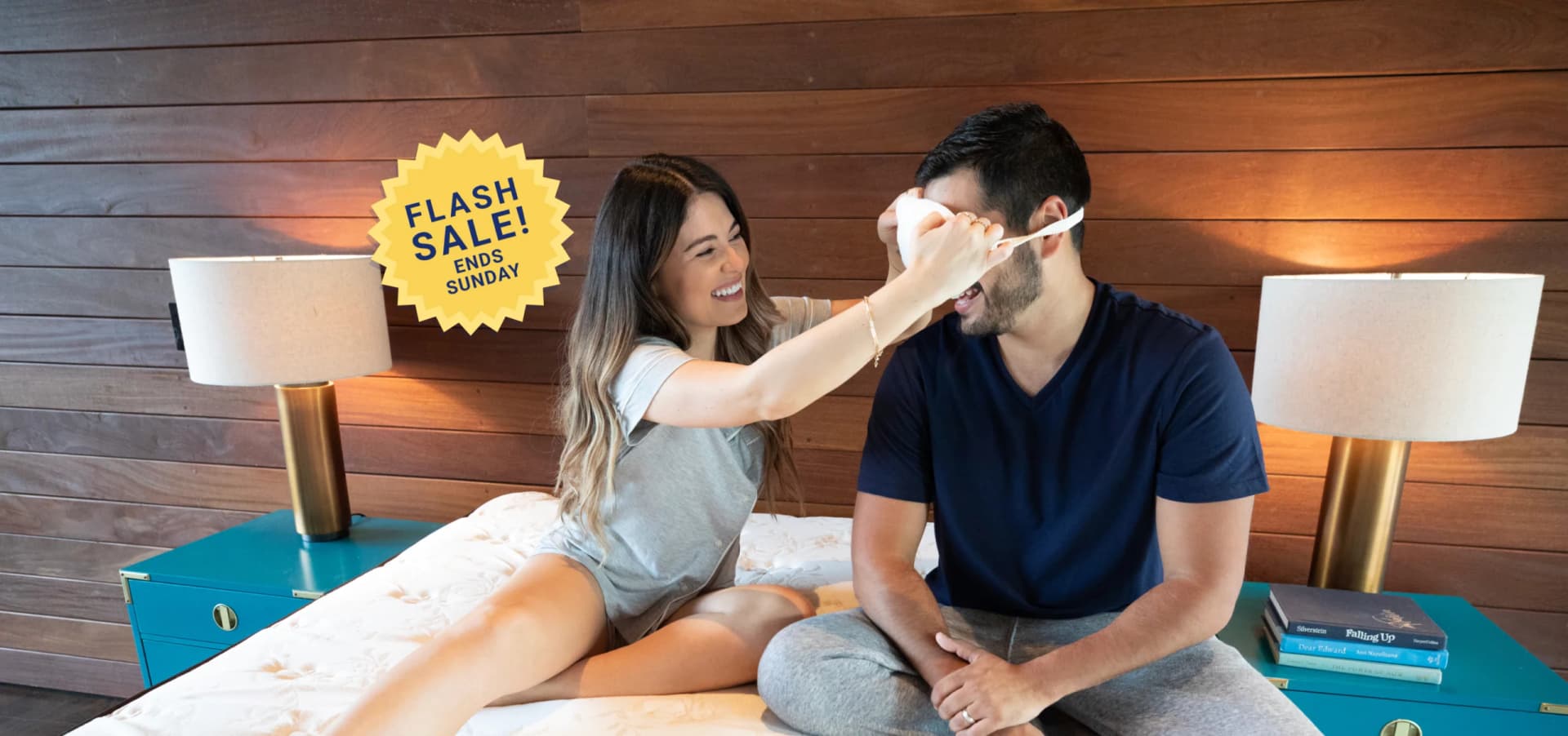 Woman puts sleep mask on male while sitting on their new mattress.