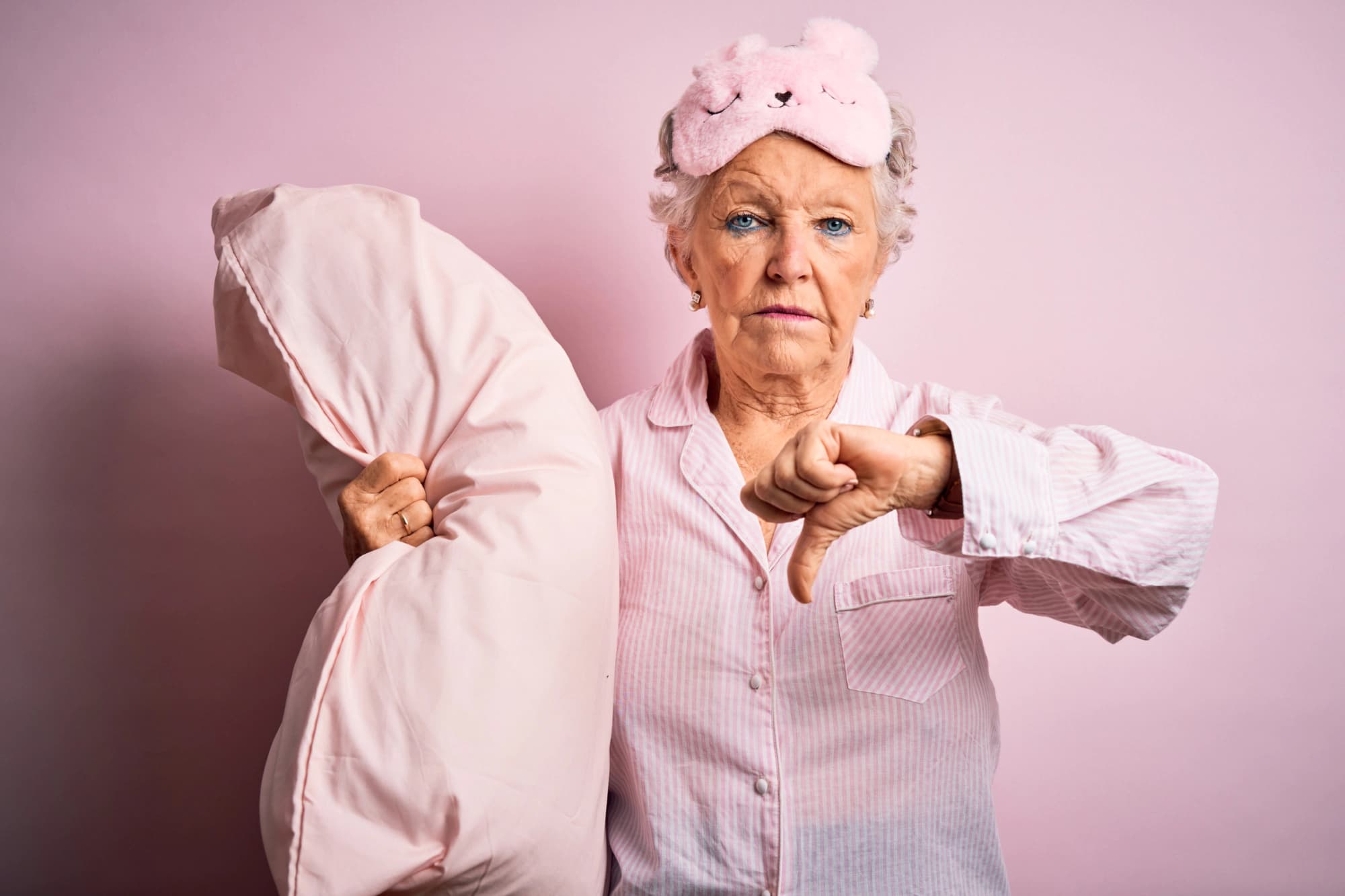 Older woman in pink pajamas holding a pillow and making a thumbs-down sign.