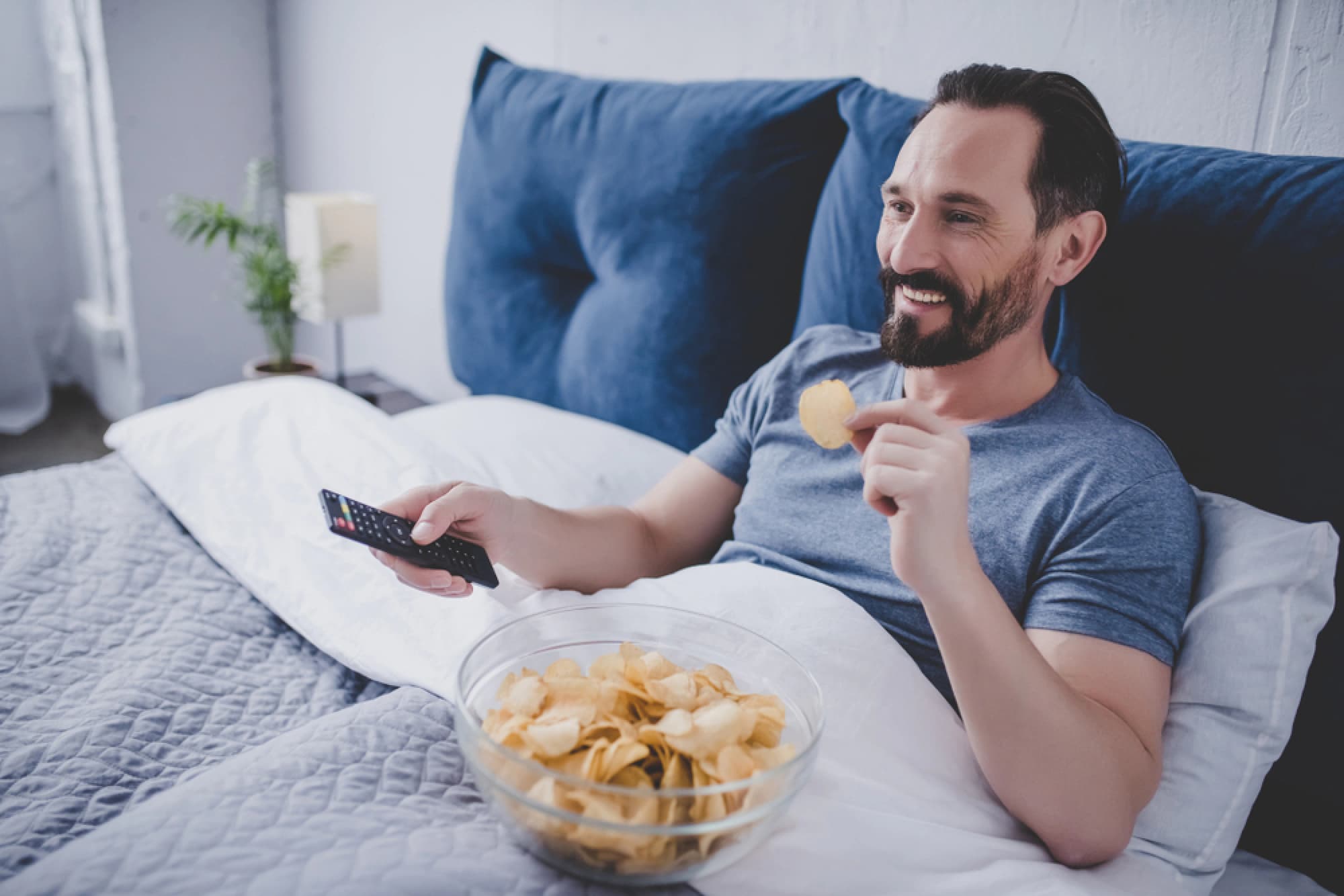 Man in bed watching TV and eating chips.