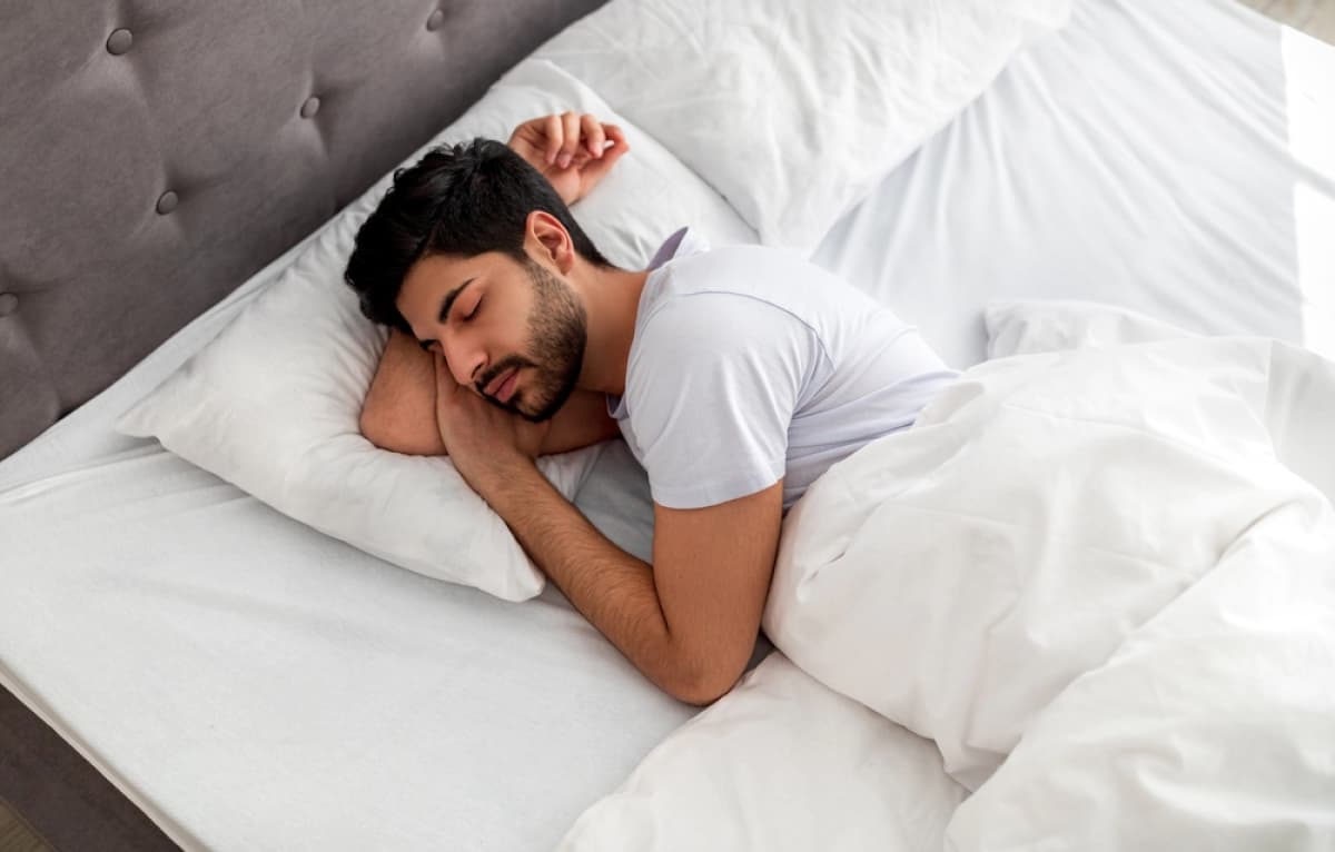 Man with brown hair asleep on a white mattress with white sheets.