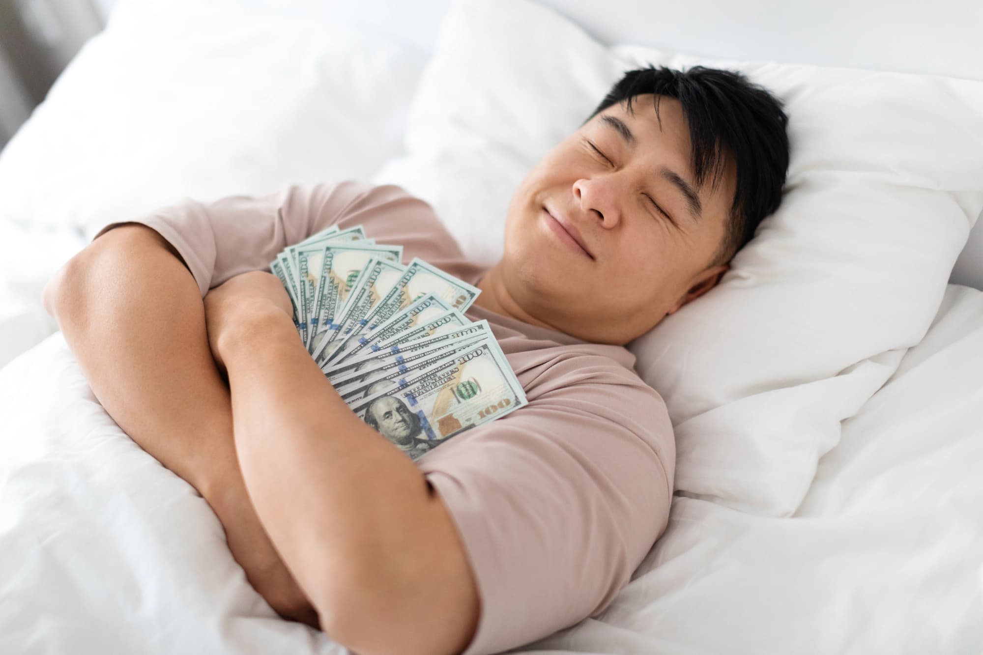 Man asleep in bed holding stacks of cash.