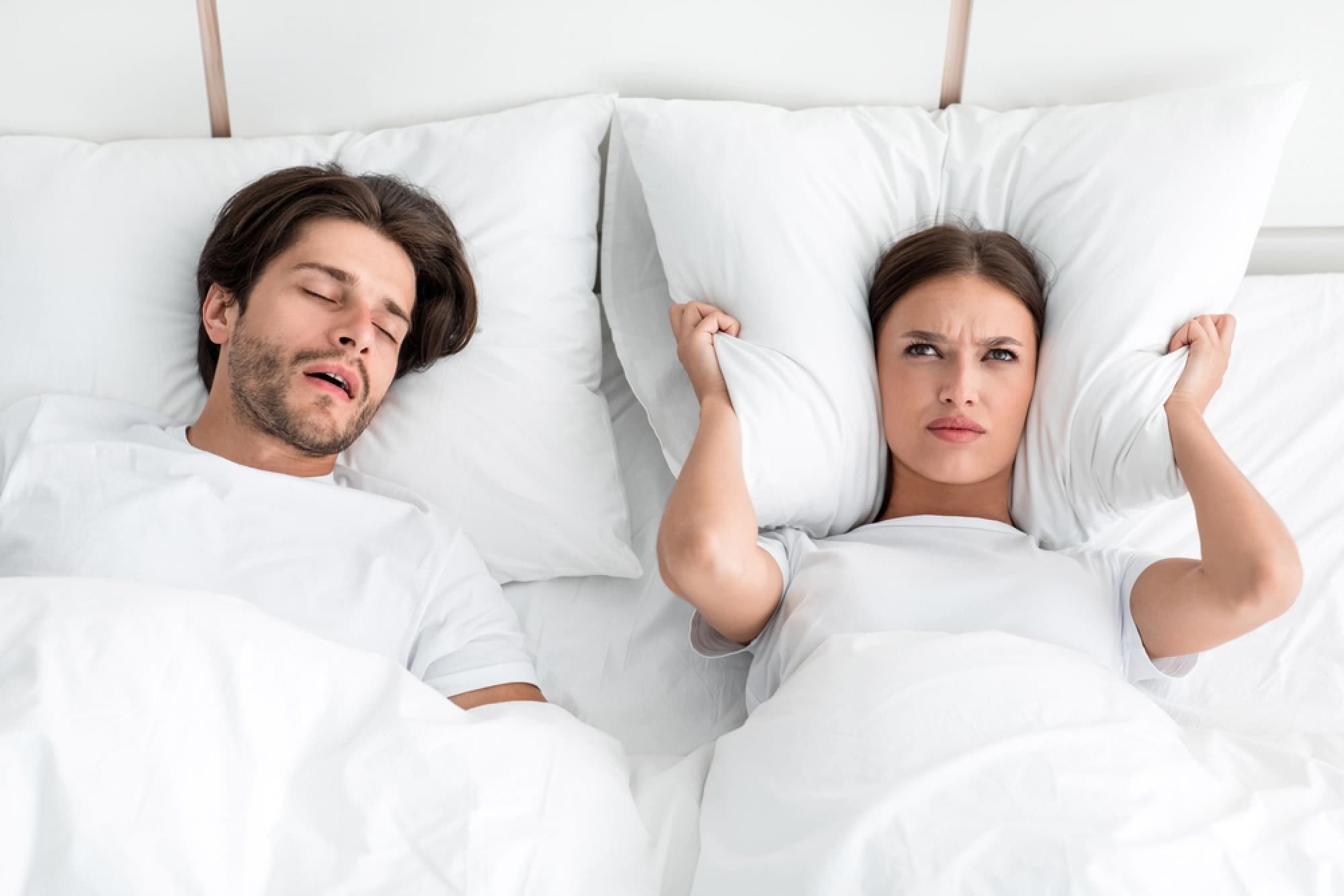 Man snoring and woman holding her pillow over her ears.