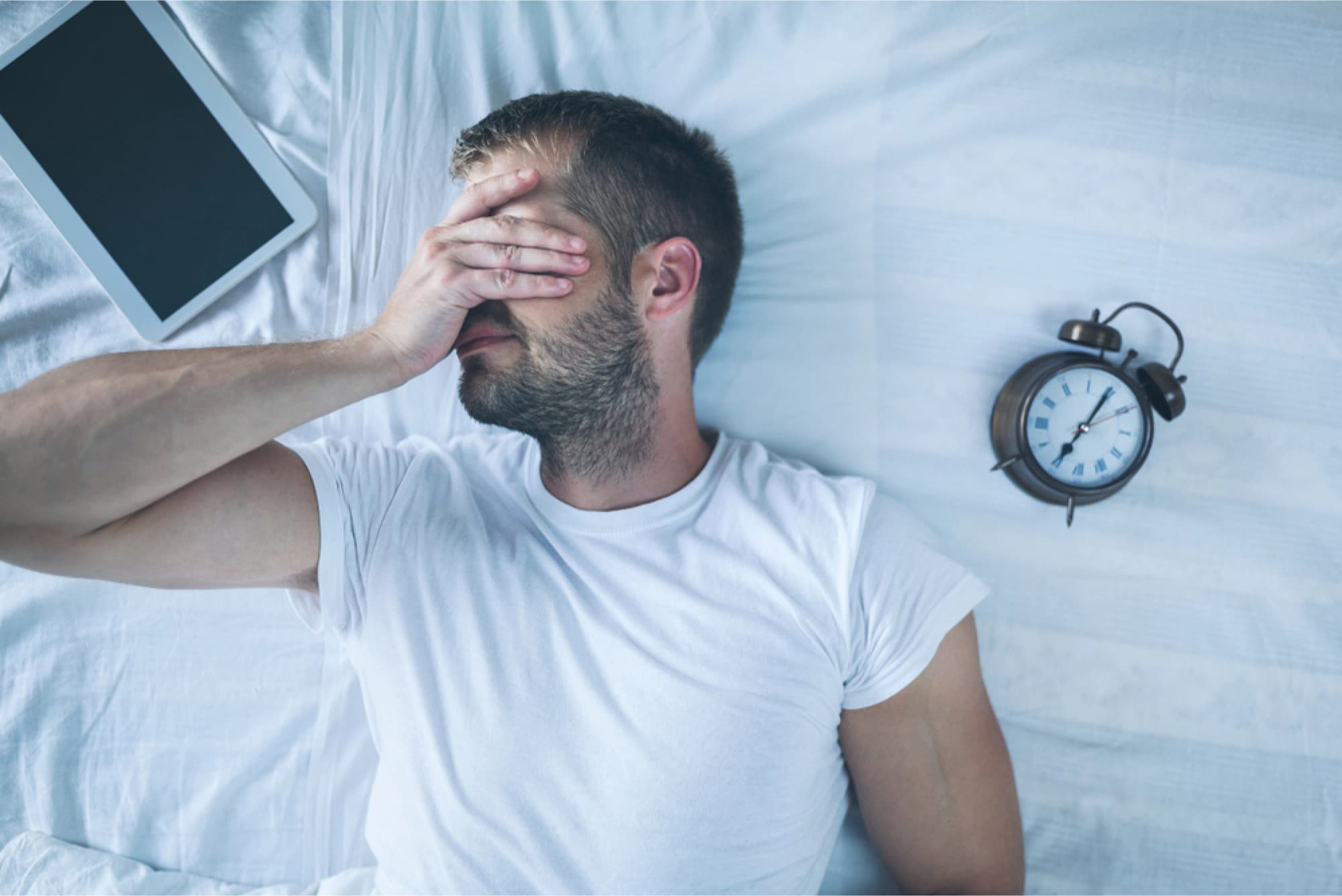 Tired man sleeping on bed with a clock and tablet.