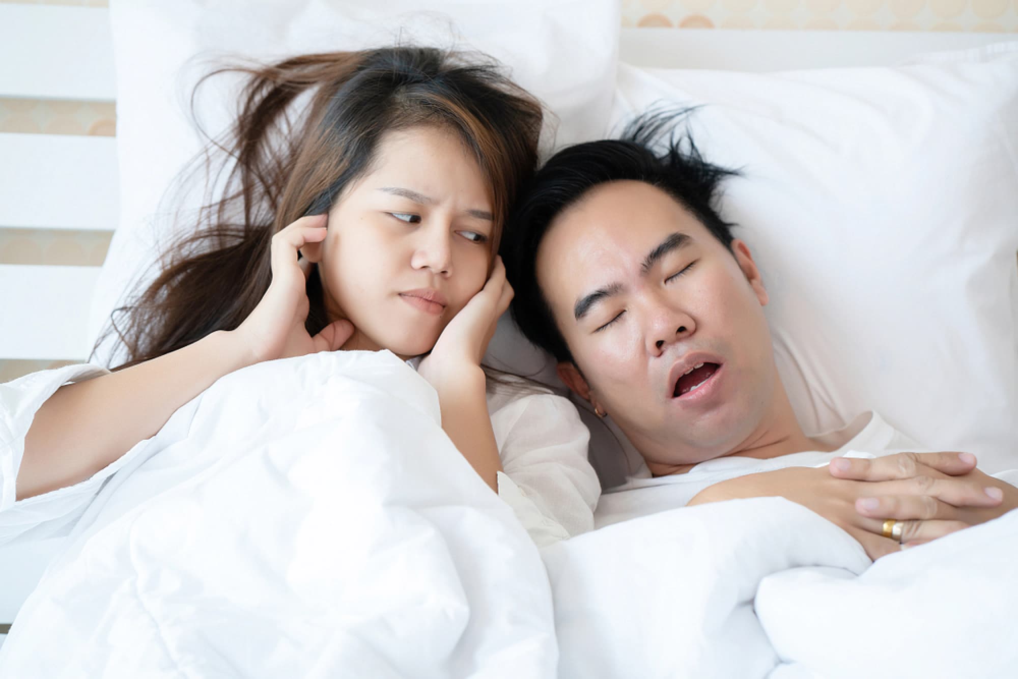 Husband snoring in bed and wife plugging her ears.